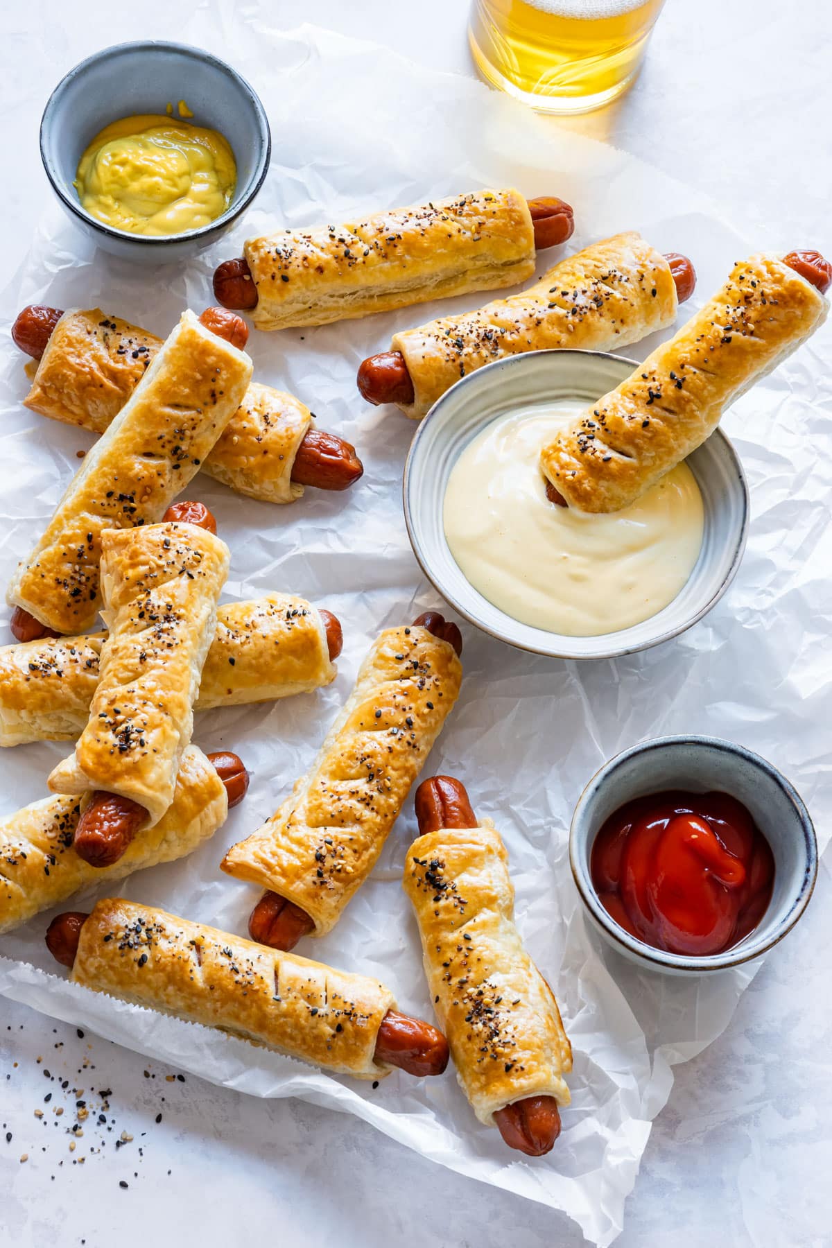 Puff pastry-wrapped hot dogs with mustard, ketchup and aioli dip on parchment paper.