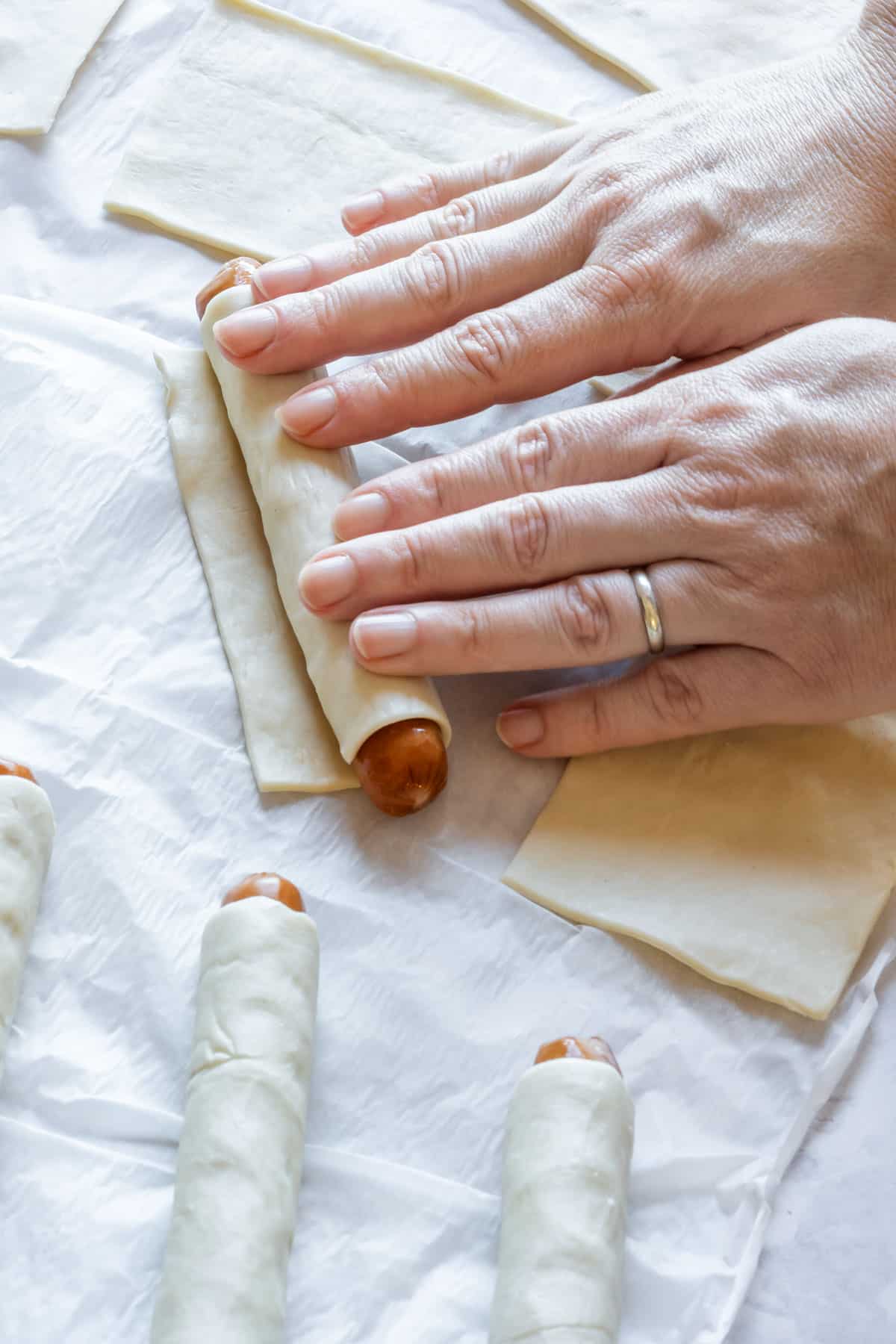 Rolling puff pastry around hot dogs. Several wrapped hot dogs on parchment paper.