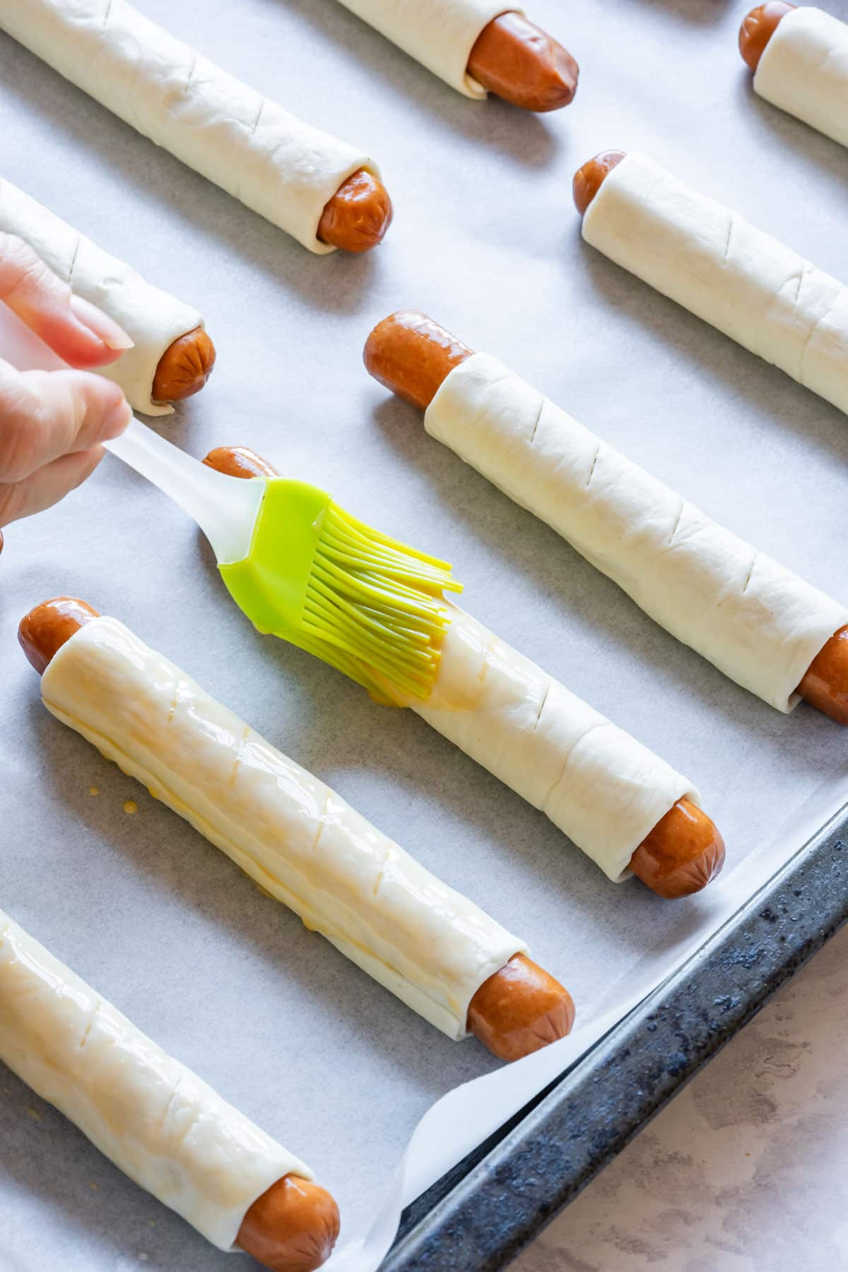Julia is brushing egg wash on puff pastry-wrapped hot dogs on a baking sheet, ready to bake.