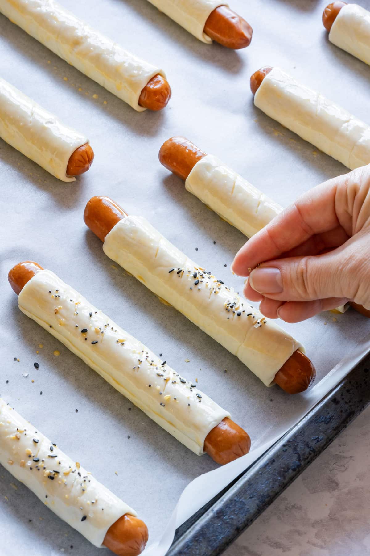 Julia sprinkling seasoning on puff pastry wrapped hot dogs on a baking sheet.