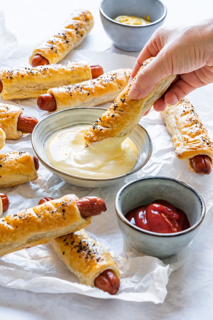 Hand dipping a puff pastry hot dog in sauce, surrounded by more puff pastry hot dogs and dipping sauces.