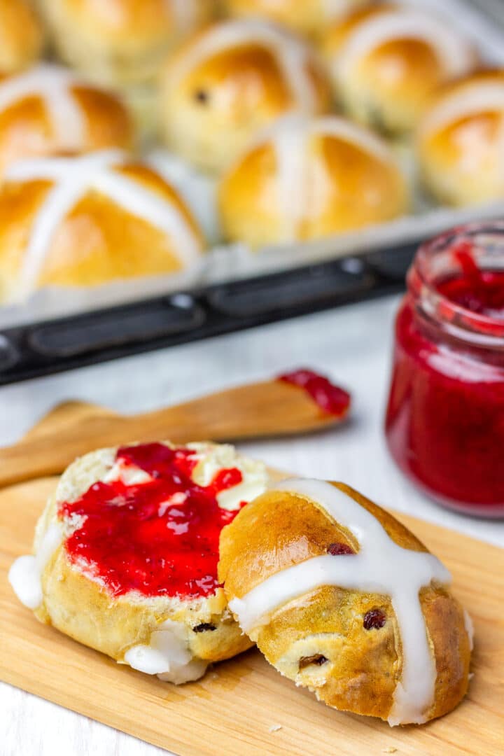 Homemade sliced hot cross bun topped with butter and jam, with a jar of strawberry jam in the background.