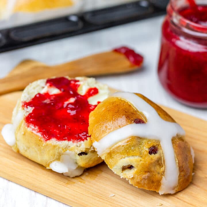 Sliced hot cross bun with butter and jam on a wooden board, with a tray of buns in the background.