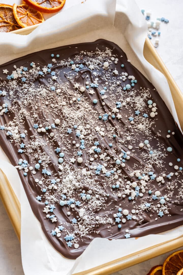 Dark chocolate bark topped with blue and white sprinkles and shredded coconut, with dried orange slices nearby.