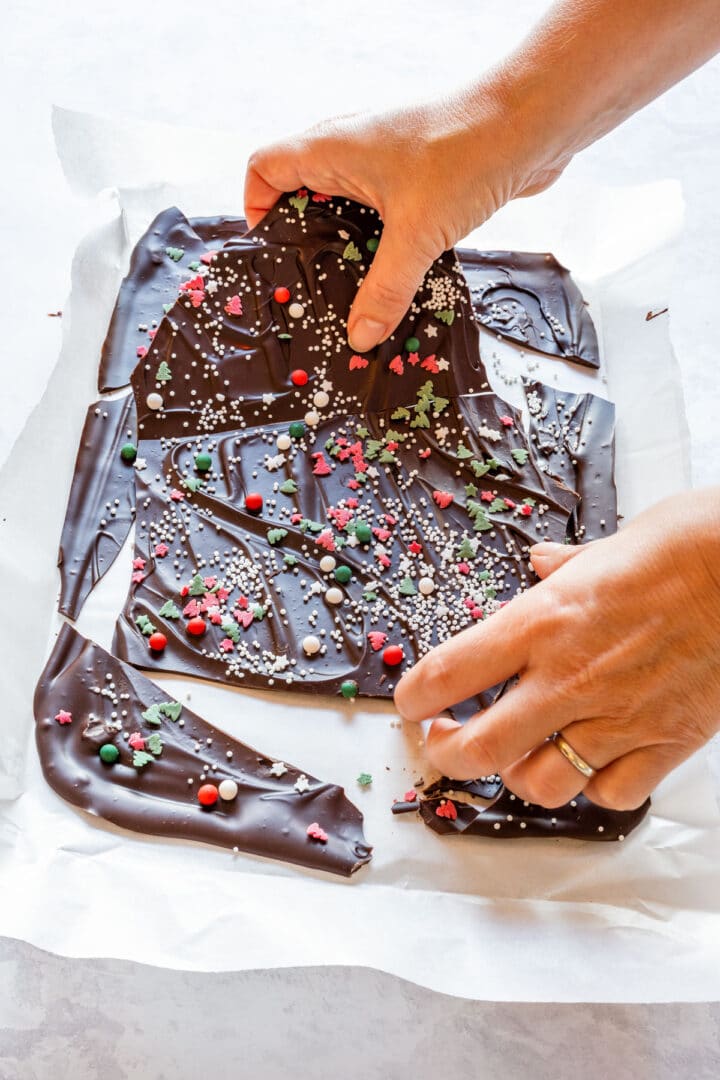 Hands breaking chocolate bark into shards, showing a glossy finish and sprinkles.