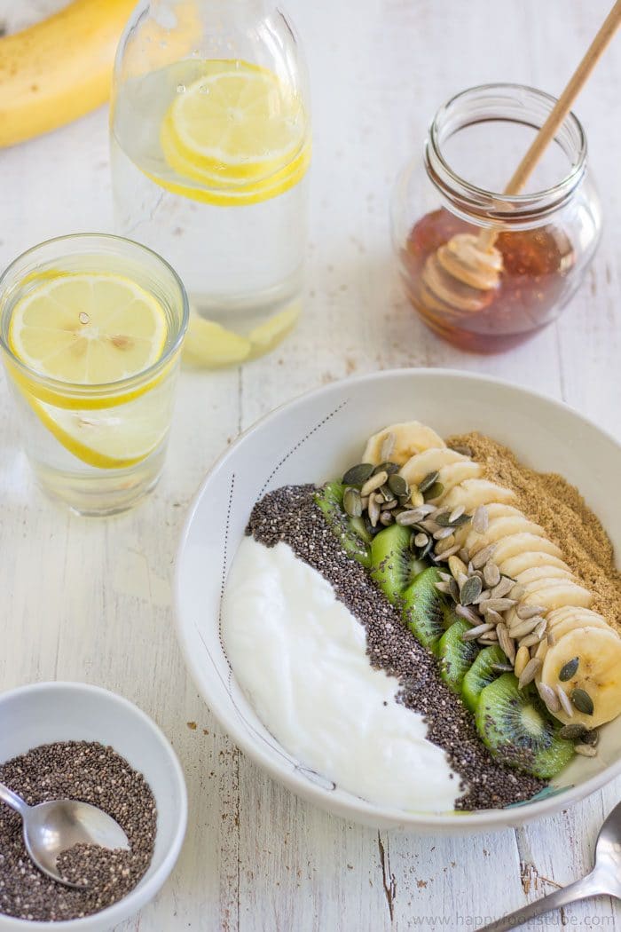 Breakfast yogurt bowl with banana, kiwi, chia and sunflower seeds next to lemon water and honey jar.
