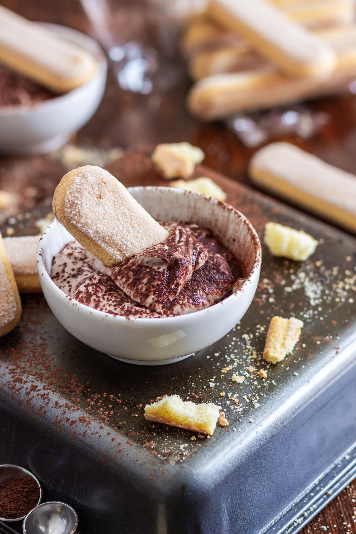 A small bowl of tiramisu dip with a ladyfinger biscuit, surrounded by more biscuits.