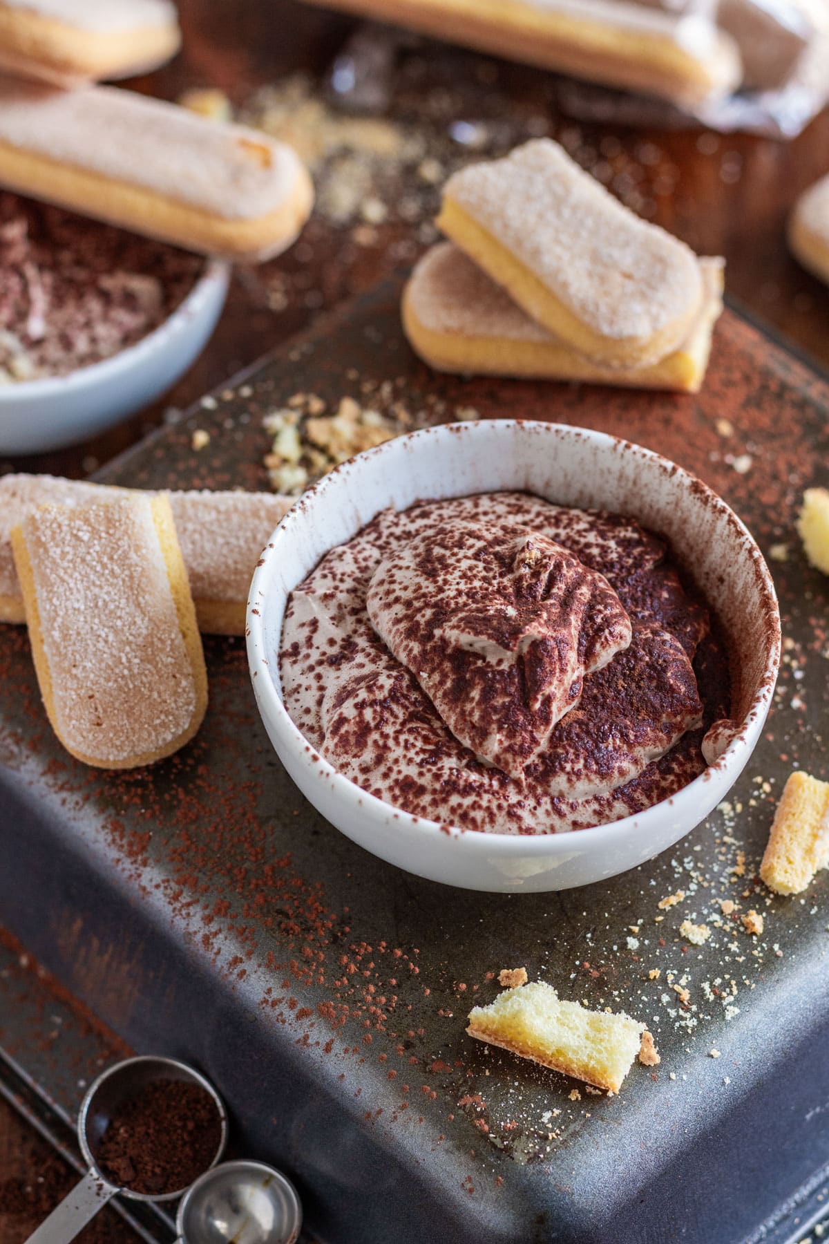 A bowl of tiramisu dessert dip with ladyfingers, cocoa powder and crumbs scattered around.