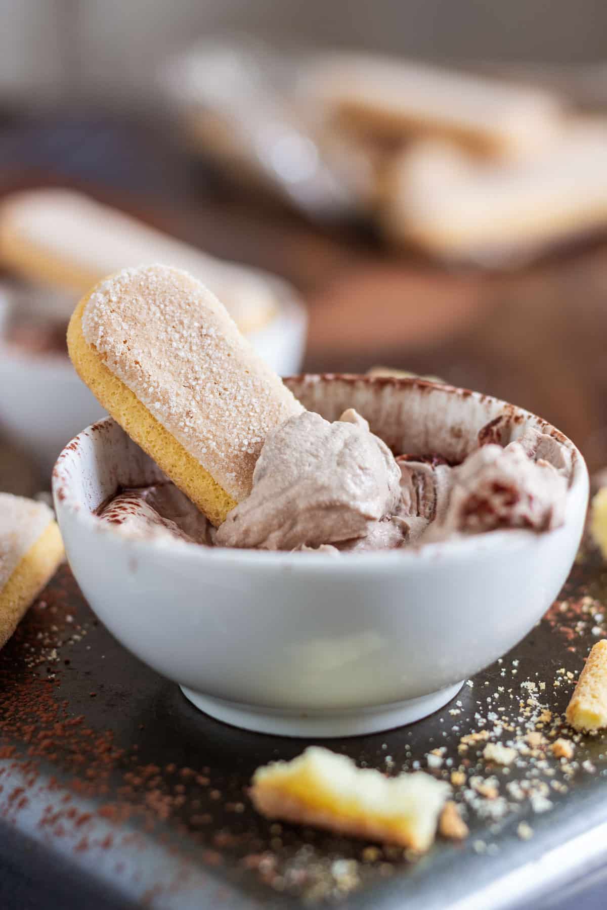 A ladyfinger dipped in coffee-flavored tiramisu dip in a white bowl, with crumbs and more ladyfingers around.