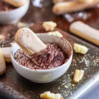 A bowl of tiramisu dip with a ladyfinger dipped in it, surrounded by more ladyfingers and crumbs.