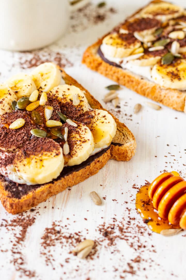 Two slices of banana toast topped with seeds, chocolate shavings and honey, with a honey dipper on the side.
