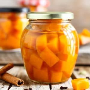 Glass jar of pickled pumpkin cubes in brine on a wooden table with a cinnamon stick.