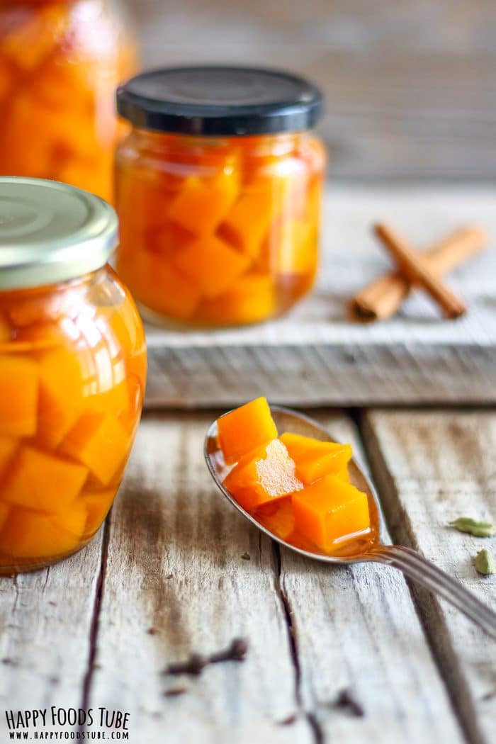 Pickled Pumpkin Images Close-up spoon with tender pumpkin cubes in brine, jar in the background with a cinnamon stick and cardamom nearby.