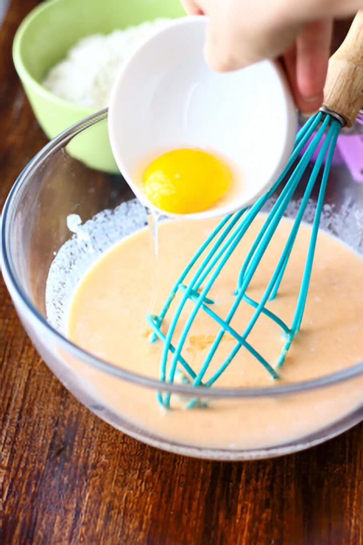 A hand is pouring an egg from a small white bowl into a mixing bowl with pancake batter and a blue whisk.