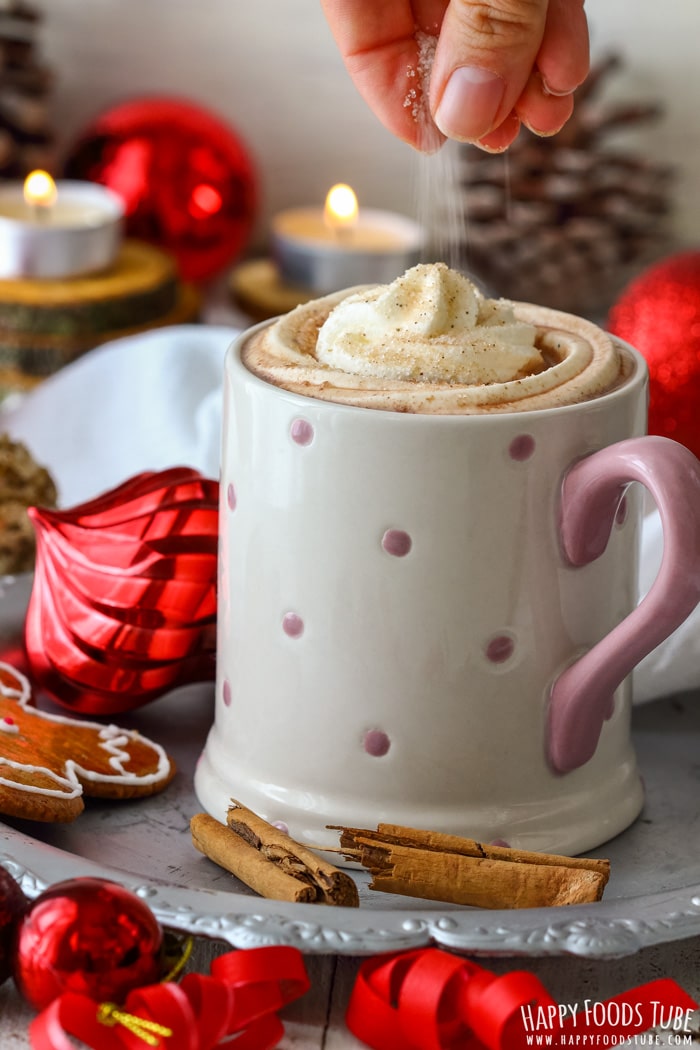 A hand sprinkles gingerbread sugar on whipped cream atop a hot chocolate in a mug.