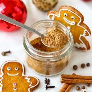 Homemade gingerbread spice mix in a small glass jar with a spoon, surrounded by gingerbread cookies, cinnamon sticks and Christmas ornaments.