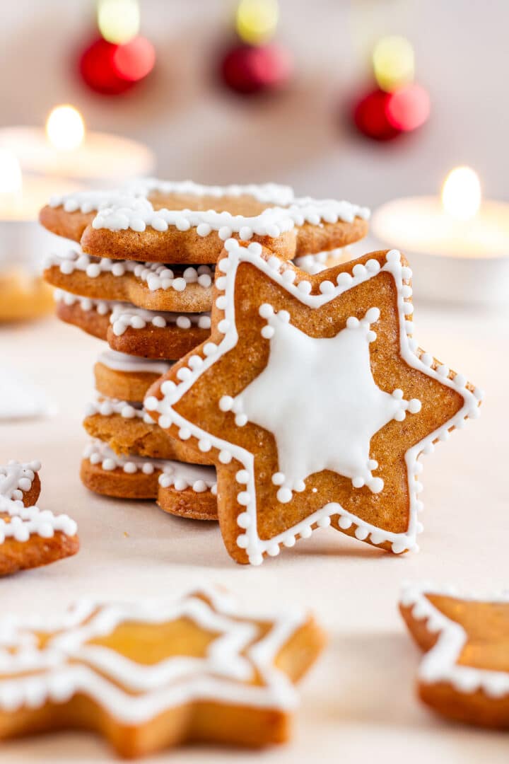 Stack of star-shaped gingerbread cookies with white royal icing, with candles and Christmas ornaments in the background.