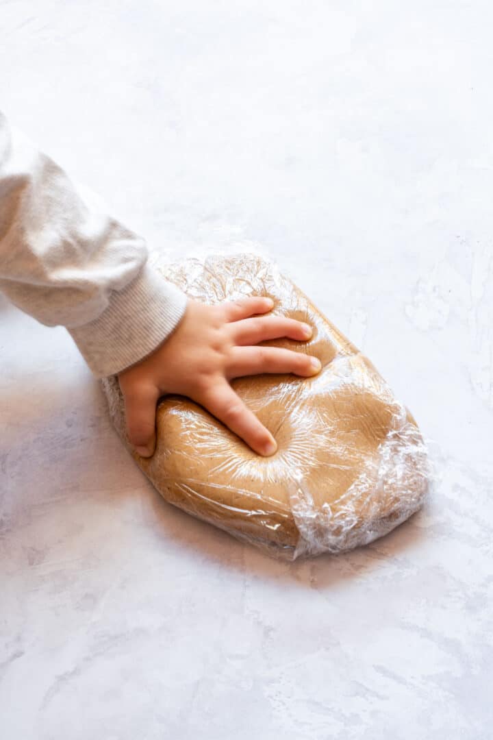 Child’s hand pressing chilled gingerbread dough wrapped in plastic on a work surface.