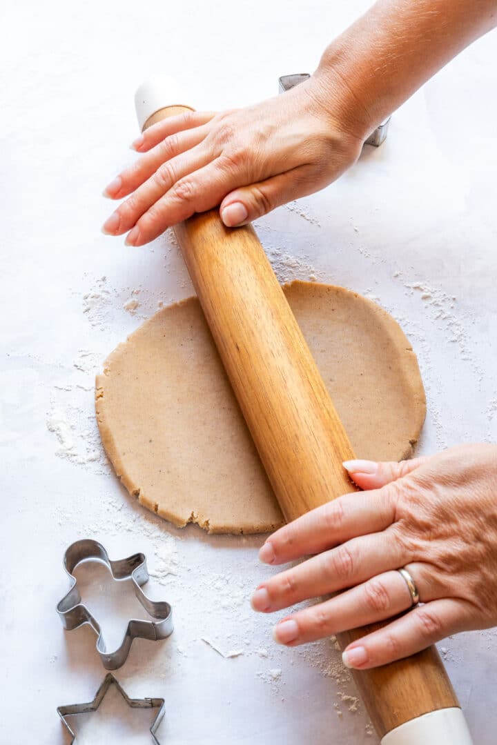 Rolling out gingerbread cookie dough on a floured surface with cutters beside the dough.