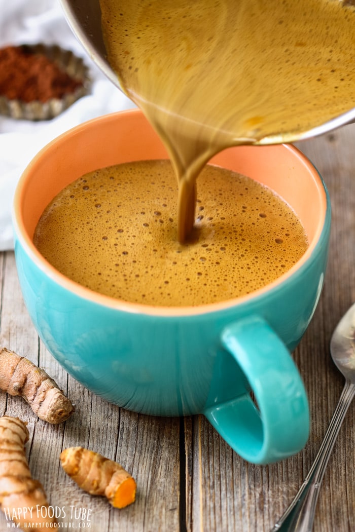 A golden turmeric hot chocolate is being poured into a turquoise mug, with turmeric roots and a spoon nearby.
