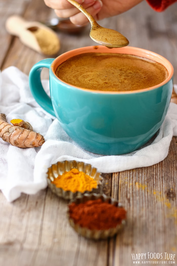 Julia holding a spoon over a large blue mug of turmeric hot chocolate, with turmeric and spices on a wooden table.