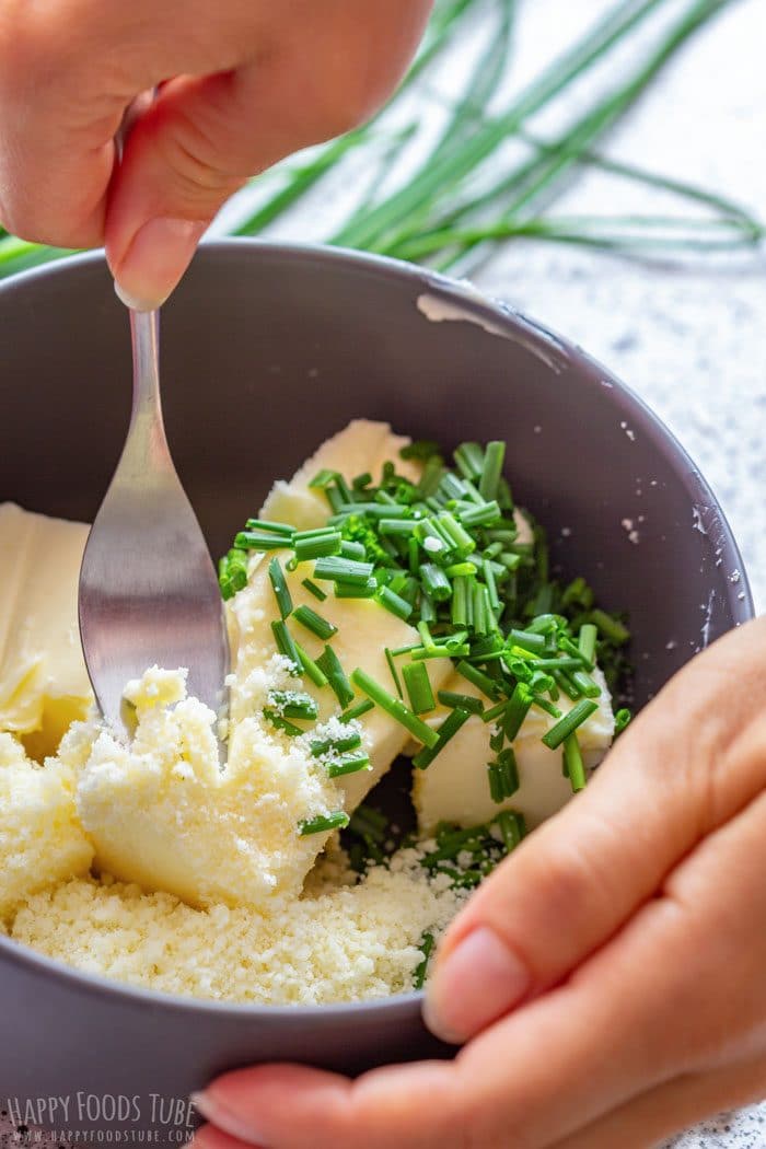 A fork blending softened butter with chopped chives and grated Parmesan in a dark bowl.