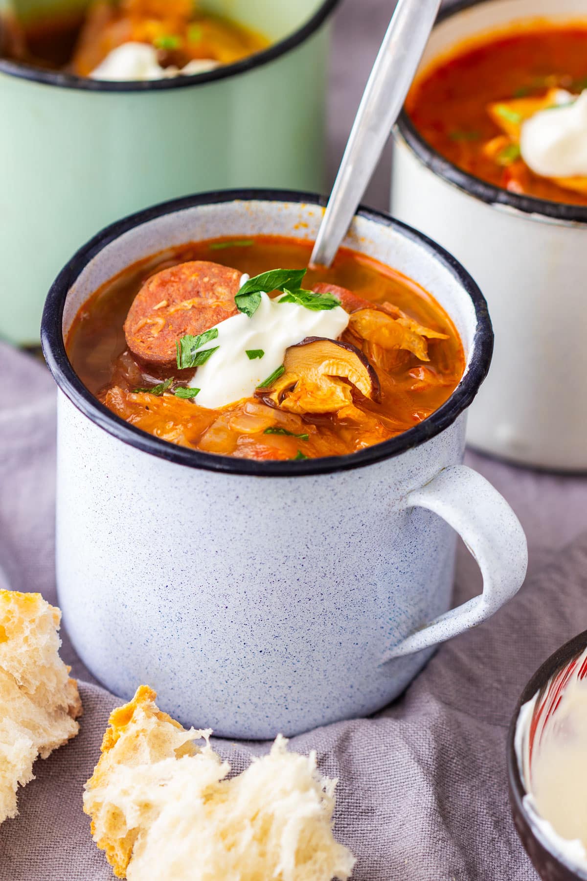 A rustic mug filled with sauerkraut soup, topped with sour cream and parsley, with bread on the side.