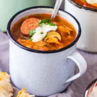 A rustic enamel mug filled with sauerkraut soup with chorizo, garnished with sour cream and parsley, served with bread.