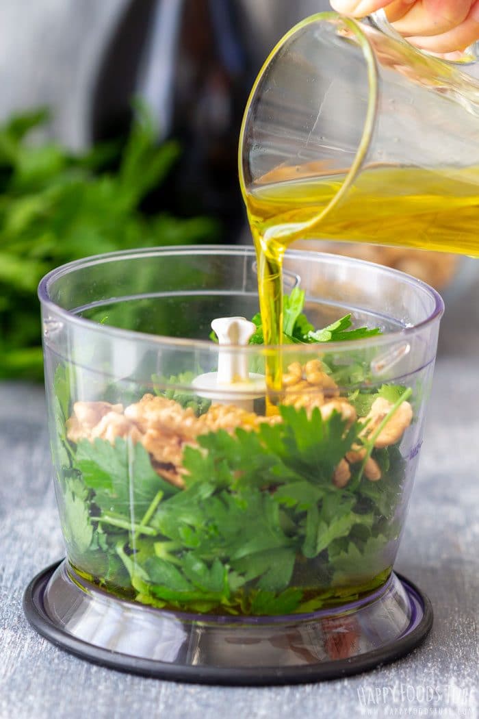 Pouring olive oil into a food processor with parsley and walnuts.