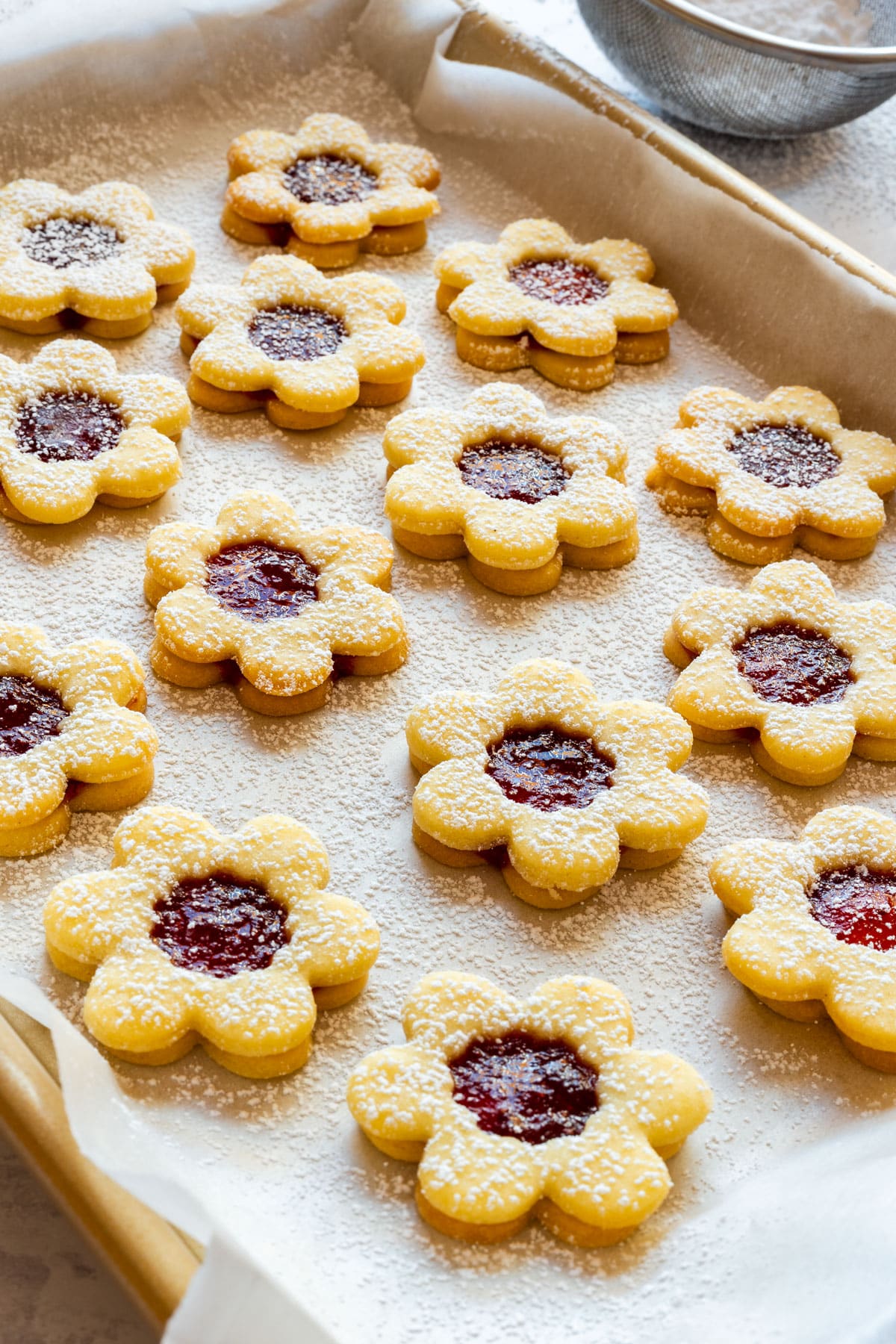 Flower shaped Linzer cookies with strawberry jam centers on a parchment lined tray, dusted with powdered sugar.