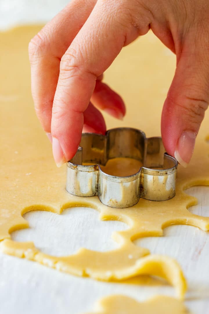 Hand pressing a flower cookie cutter into rolled Linzer dough on a floured work surface.