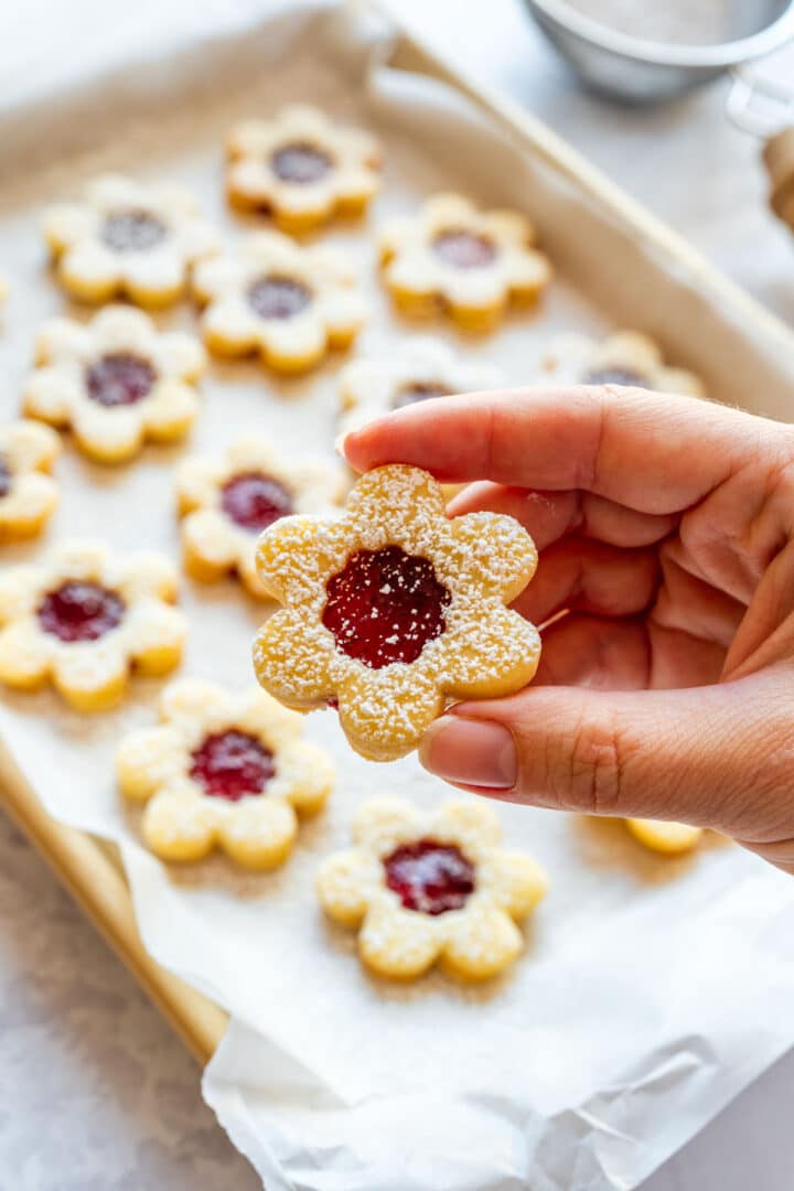 Hand holding one small flower Linzer cookie over a tray of more cookies in the background.