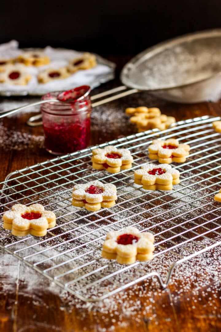 Linzer cookies with raspberry jam cooling on a wire rack with a jar of jam and powdered sugar nearby.