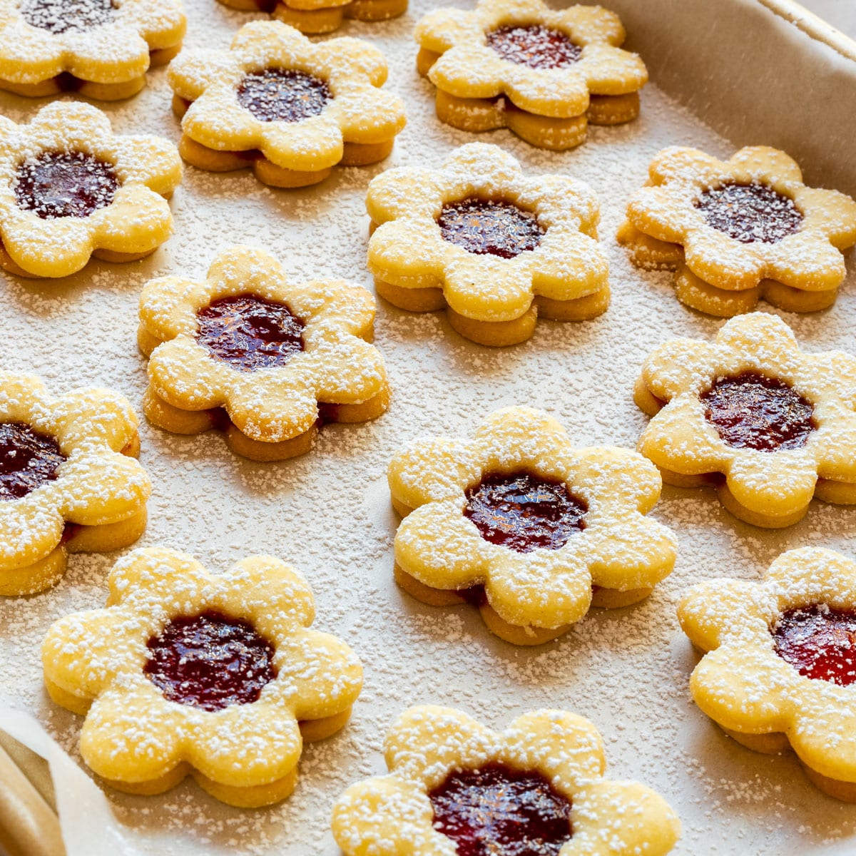 Tray of small flower Linzer cookies with strawberry jam centers, generously dusted with powdered sugar.