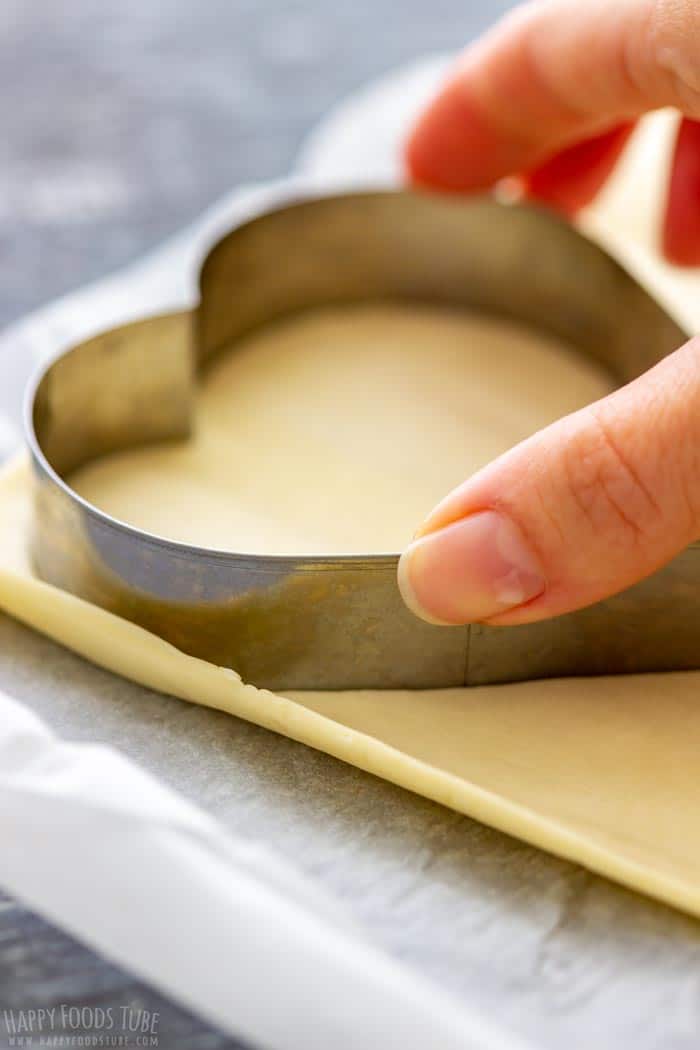 A hand is pressing a heart-shaped cookie cutter into a sheet of puff pastry on a parchment-lined surface.