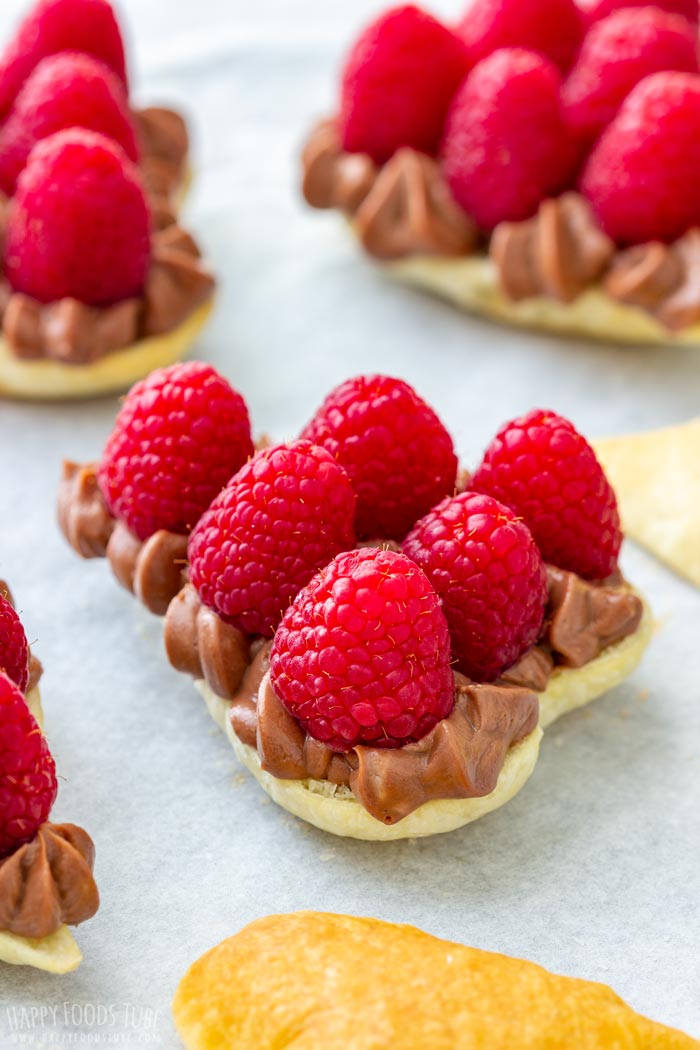 Heart-shaped puff pastry topped with chocolate filling and raspberries on parchment paper.