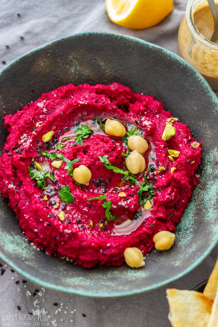 A vibrant beet hummus garnished with chickpeas, herbs and seeds in a rustic bowl, with lemon and tahini nearby.