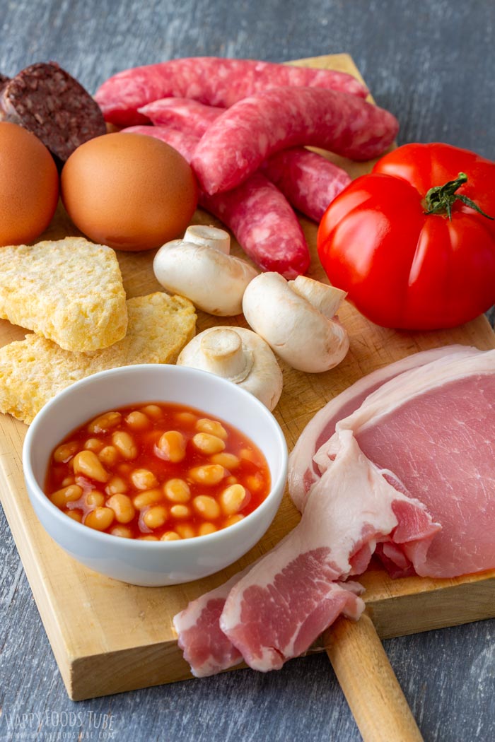 Irish breakfast ingredients on a wooden board, including bacon rashers, sausages, black pudding, eggs, baked beans, tomatoes, mushrooms and hash browns.
