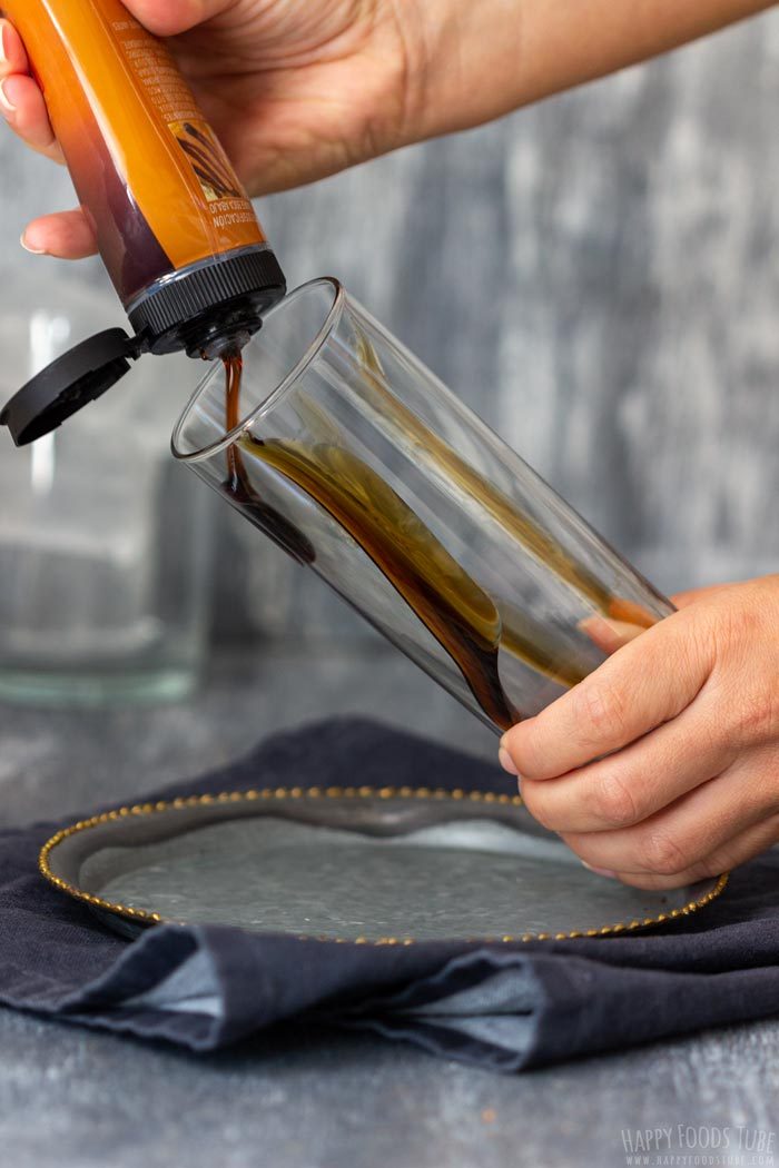 Squeezing pourable caramel sauce around the inside of a tall glass.
