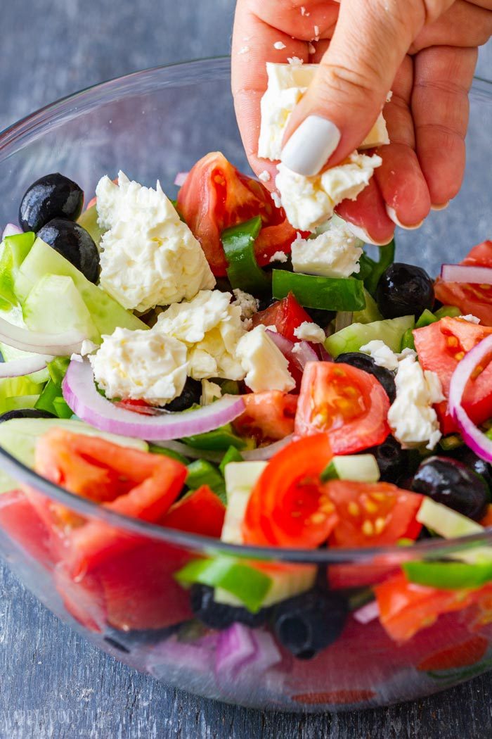 A hand adding crumbled feta to tomatoes, cucumber, green pepper, red onion and black olives.