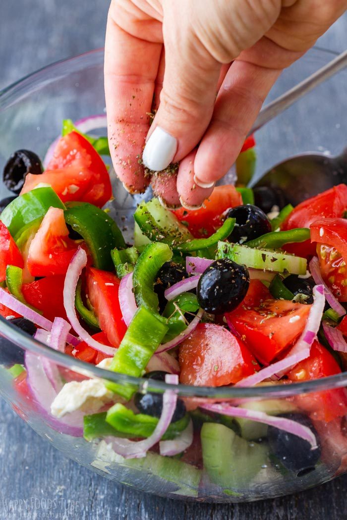 A hand sprinkling dried oregano over tomatoes, cucumber, green pepper, red onion and olives.