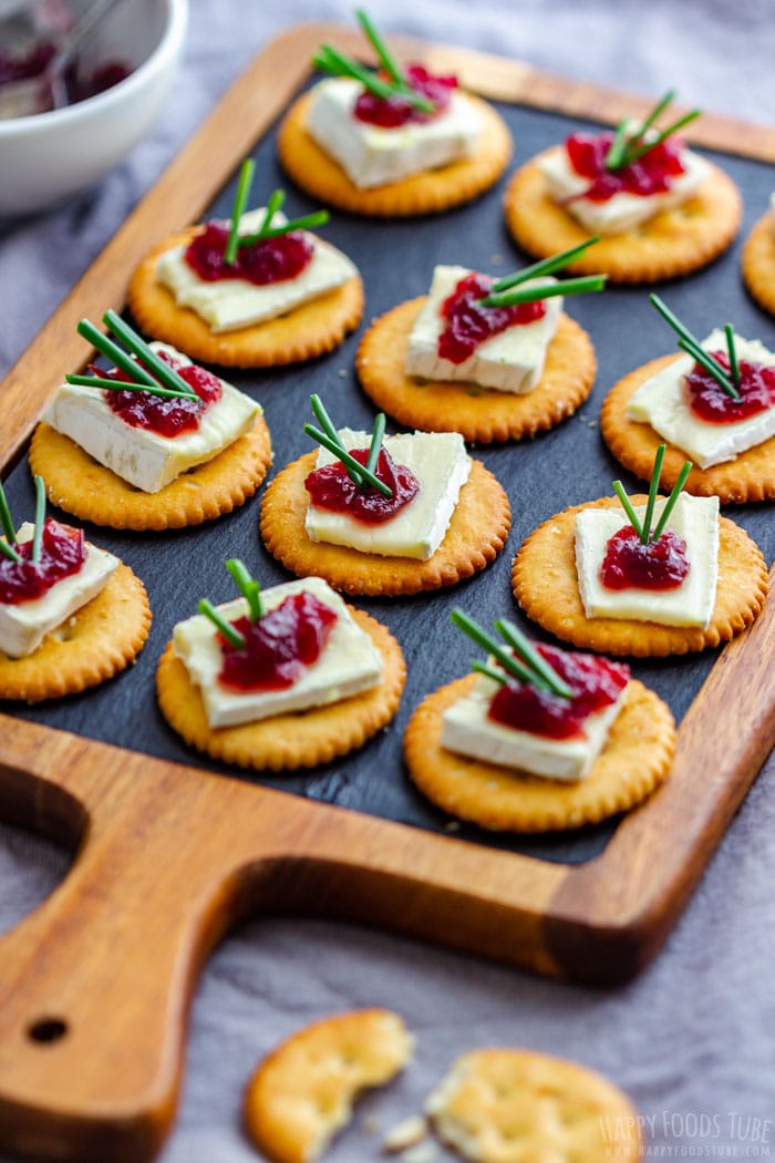 Serving tray with round crackers topped with brie, a dollop of cranberry jam and garnished with green chives.