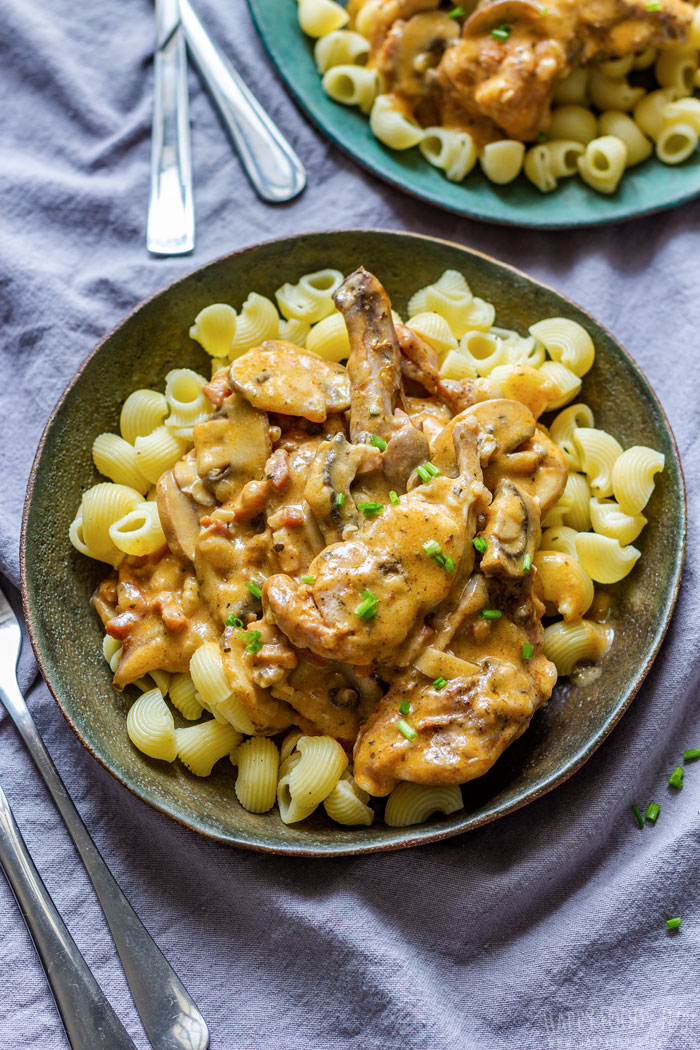 A bowl of pasta with creamy rabbit and mushroom sauce, garnished with chives, on a table setting.