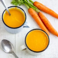 Two mugs of carrot ginger soup with fresh carrots and a spoon on a white table.