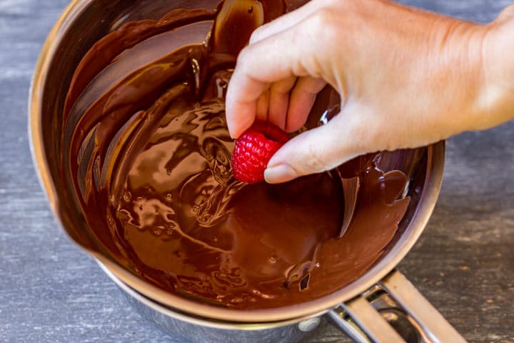 Hand dipping a raspberry into melted chocolate.