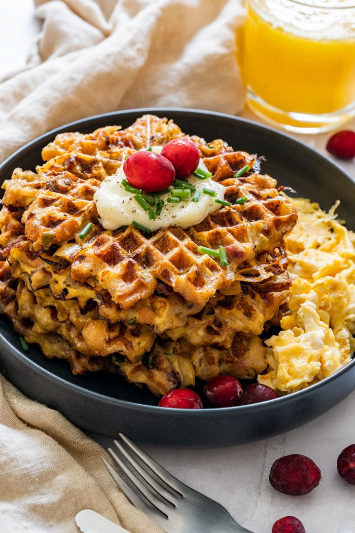 A stack of Thanksgiving leftover stuffing waffles topped with garlic sauce, chives and cranberries, served with scrambled eggs.