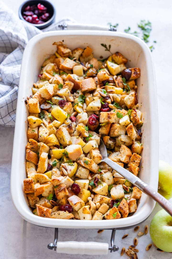 Overhead of golden bread cubes with apples and cranberries, spoon resting in pan.