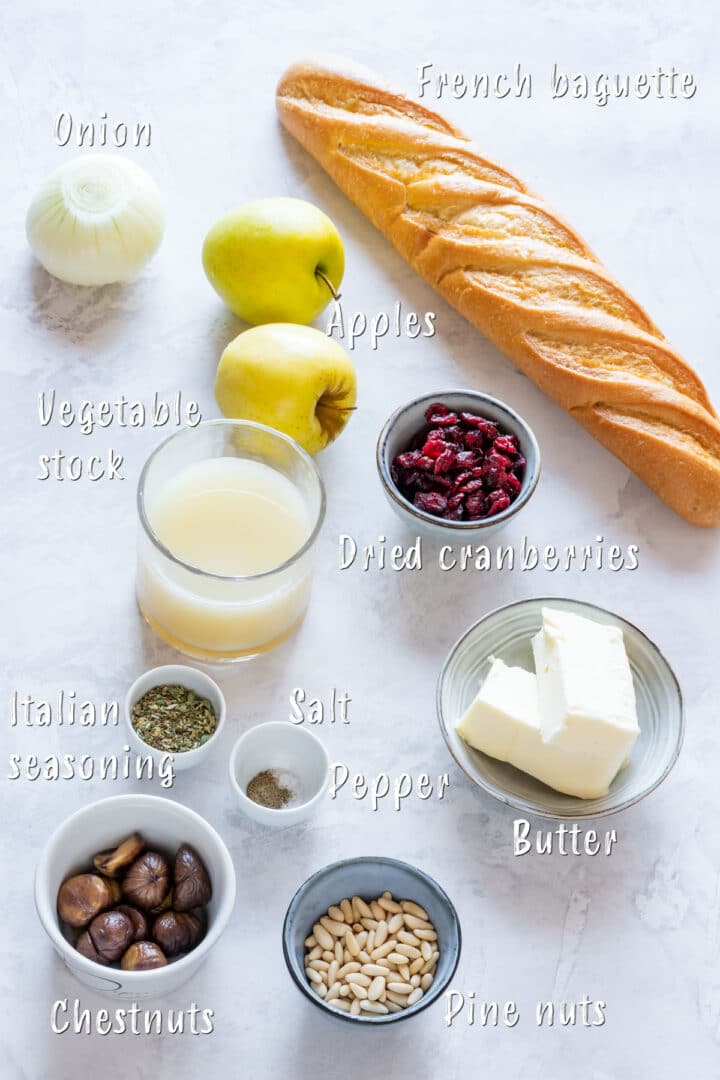 Ingredient lineup with baguette, apples, dried cranberries, stock, herbs, butter, chestnuts, pine nuts and onion.