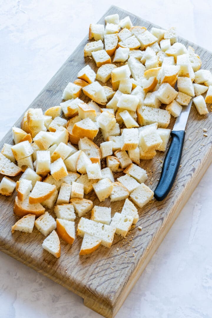 Baguette cut into cubes on a wooden board with a knife.