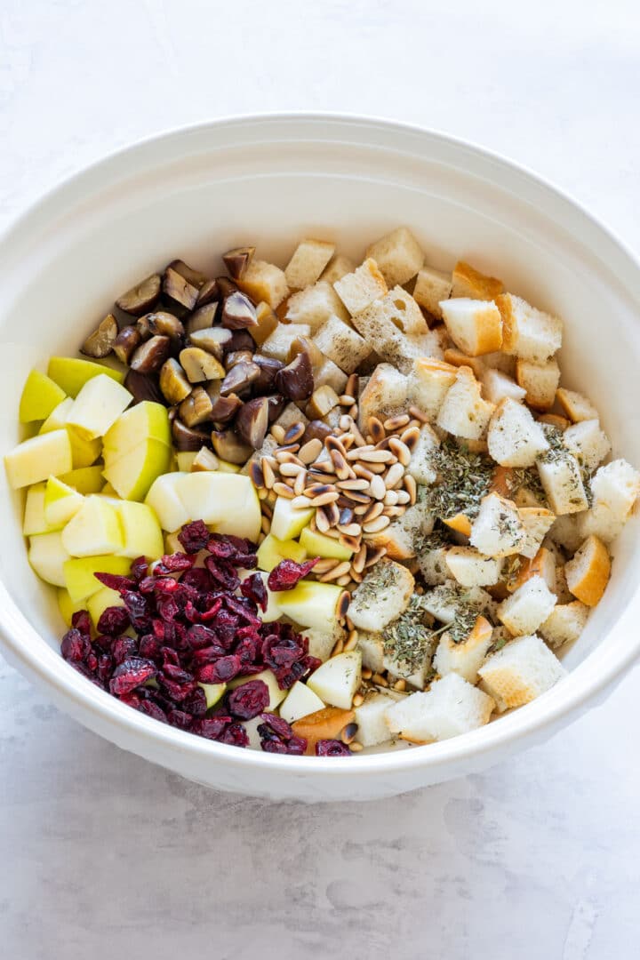 Unmixed bowl showing piles of bread, apples, cranberries, chestnuts, pine nuts and herbs.